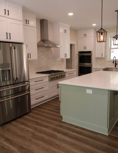 Modern kitchen with white cabinets, stainless steel appliances, wooden floor, and a green island with a white countertop. Two pendant lights hang above the island.