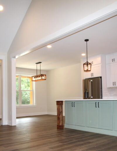 Modern kitchen and living area with wooden flooring, a large kitchen island with pendant lights, white cabinets, and a stainless steel range hood. Dark front door visible in the background.