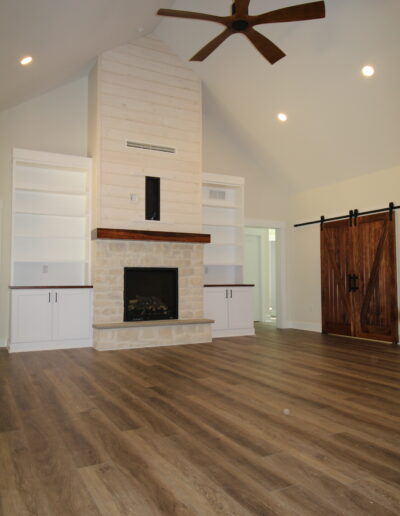 Spacious living room with wooden flooring, a stone fireplace, built-in shelves, ceiling fan, and sliding barn door.