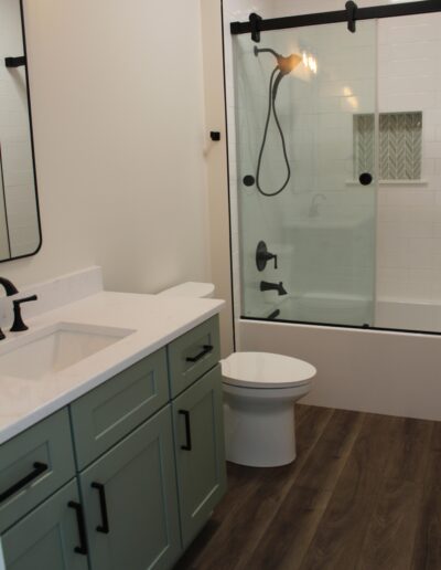 A bathroom with a green vanity, white countertop, mirror, toilet, and a shower-bath with a glass sliding door. The floor is wood-textured.