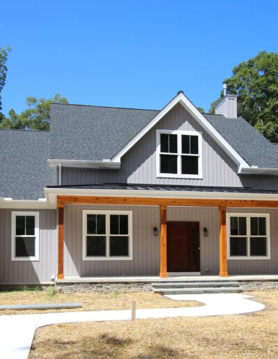 A modern, two-story house with a grey facade and a porch, set amidst a backdrop of trees under a clear blue sky.