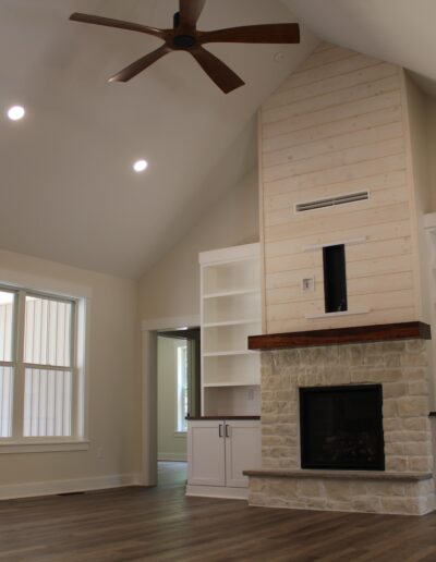 A spacious living room with a vaulted ceiling, ceiling fan, stone fireplace, built-in shelves, and hardwood flooring. Large windows provide natural light.