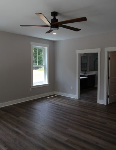 A modern, empty room with dark wood flooring, gray walls, two large windows, a ceiling fan, and doorways leading to an adjacent room.
