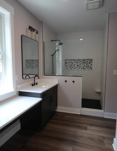 Modern bathroom with dark vanity, large mirror, and a walk-in shower featuring a decorative tile niche. Wood flooring and natural light from a window are visible.