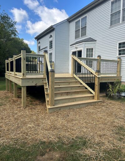 A wooden deck with stairs leading to a two-story white house. The deck features black railings and is surrounded by a grass yard with some straw patches.