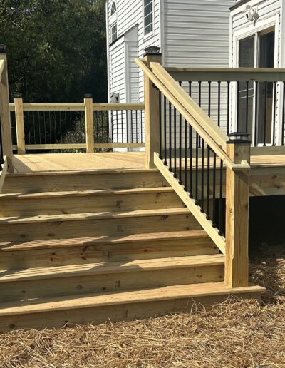 A new wooden deck with black railing is attached to a white house. It has steps leading down to a mulched area with a few plants.