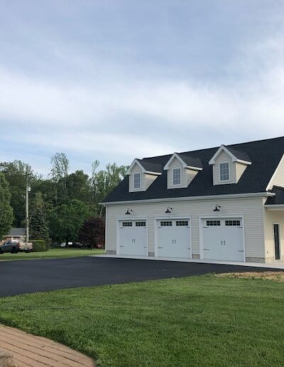 A white detached garage with three doors, dormer windows, and a covered side porch stands near a blacktop driveway and adjacent green lawn.