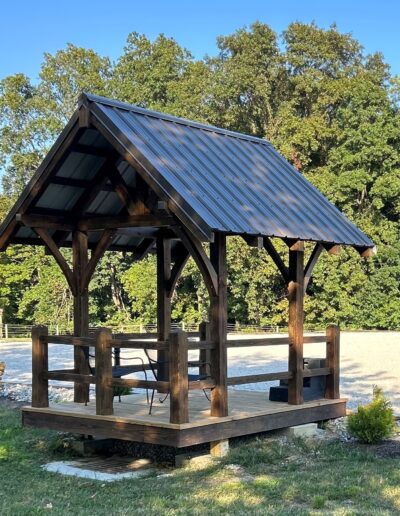 A small wooden gazebo with a metal roof stands in a grassy area, surrounded by trees. A chair is placed inside.