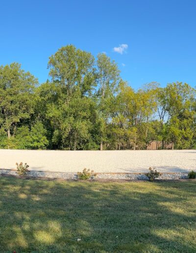 A large outdoor riding arena with a sand surface, bordered by trees and grass, under a clear blue sky.